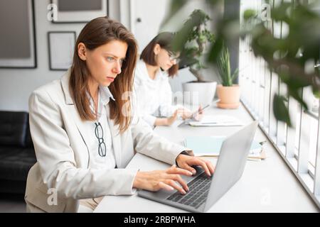 Responsable de bureau féminin travaillant sur ordinateur portable dans l'espace de coworking, collègue utilisant la tablette numérique assis au bureau sur le fond Banque D'Images