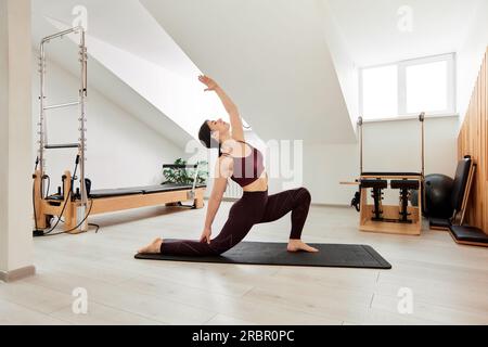 Jeune fille fait Pilates dans un studio lumineux sur un tapis de yoga. Mince brunette en body bordeaux fait un stand pour entraîner l'équilibre et renforcer le muscl Banque D'Images