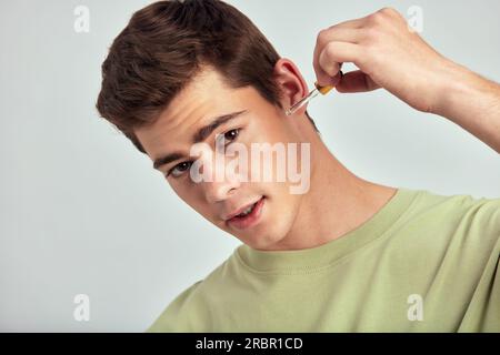 Jeune beau mec souriant avec les cheveux bruns applique le sérum de beauté sur son visage pour garder sa peau douce et lisse. Homme caucasien maintenant son être naturel Banque D'Images