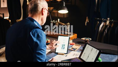 Couturier dessinant des croquis d'articles avec tablette et ordinateur portable, travaillant sur la ligne de vêtements de luxe dans l'atelier de couture. Créateur de mode fabriquant des vêtements avec des outils d'atelier professionnels. Prise de vue à main levée. Banque D'Images