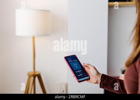 Jeune femme utilisant l'application de maison intelligente sur le téléphone cellulaire à la maison Banque D'Images