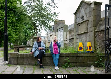 27 juin 2023.Aberdeen, Écosse. Voici deux femmes assises sur un mur dans un cimetière, utilisées comme espace public, fumant une cigarette chacune. Banque D'Images