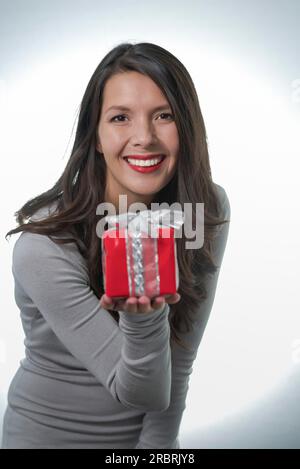 Jolie jeune femme aux longs cheveux brun et un beau sourire doux qui fait un cadeau rouge coloré pour un être cher Banque D'Images