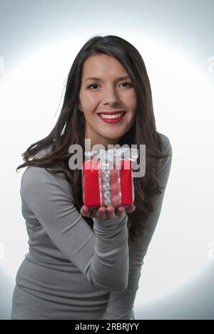 Jolie jeune femme aux longs cheveux brun et un beau sourire doux qui fait un cadeau rouge coloré pour un être cher Banque D'Images