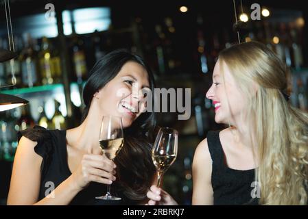 Deux bonnes amies heureuses, une blonde et une brune, grillant avec du champagne ou du vin blanc dans une discothèque branchée Banque D'Images