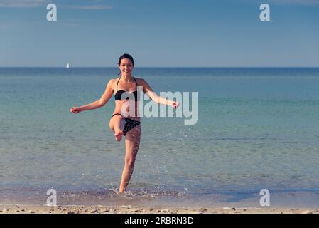 Heureuse jeune femme attirante dans un bikini batillant sur une plage tropicale s'amusant à rire et botter le jet de la mer avec son pied, avec Banque D'Images