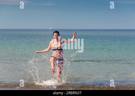 Heureuse jeune femme attirante dans un bikini batillant sur une plage tropicale s'amusant à rire et botter le jet de la mer avec son pied, avec Banque D'Images