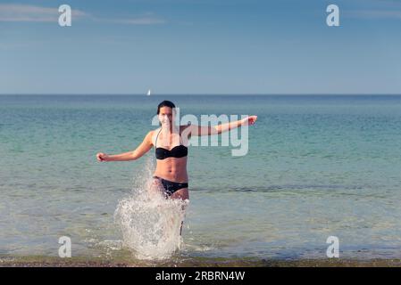 Heureuse jeune femme attirante dans un bikini batillant sur une plage tropicale s'amusant à rire et botter le jet de la mer avec son pied, avec Banque D'Images