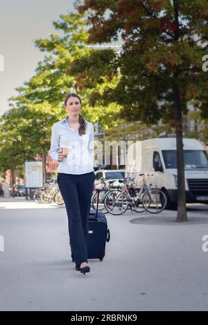 Attrayante jeune femme touriste tirant un chariot dans la rue sur ses vacances d'été avec des vélos stationnés et des voitures derrière Banque D'Images