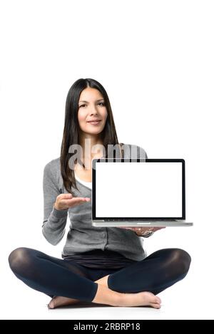 Jeune jolie femme assise sur le sol avec des jambes croisées Publicité tout nouvel ordinateur portable à portée de main. Isolé sur fond blanc Banque D'Images