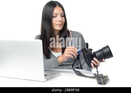 Photographe femme insérant ou retirant une carte mémoire dans son appareil photo reflex numérique professionnel alors qu'elle est assise à un bureau avec son ordinateur portable prêt à être transféré Banque D'Images