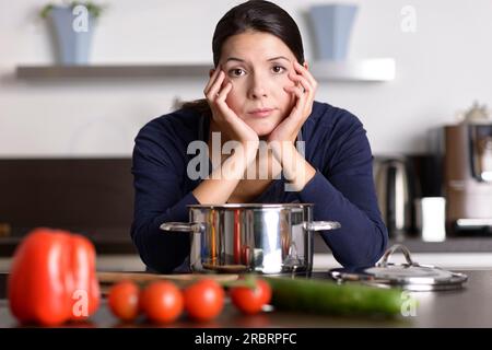 Jeune femme séduisante peu motivée préparant le dîner appuyé sur la table de cuisson regardant la caméra avec une expression de glum apathique comme elle se tient en elle Banque D'Images