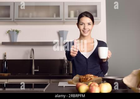 Heureuse jeune cuisinière attirante donnant un pouce vers le haut de l'approbation et du succès comme elle se tient dans sa cuisine en buvant du café avec une pomme fraîchement cuite Banque D'Images