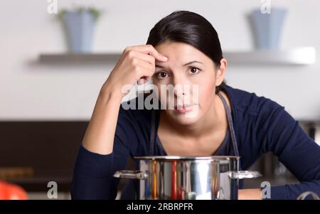 Jeune femme séduisante peu motivée préparant le dîner appuyé sur la table de cuisson regardant la caméra avec une expression de glum apathique comme elle se tient en elle Banque D'Images