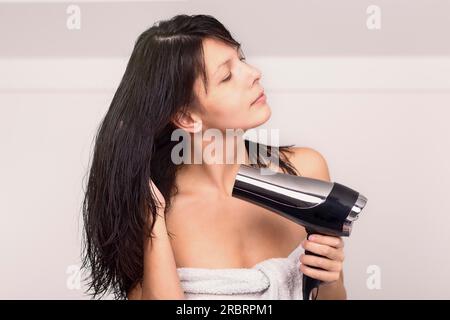 Jolie femme dans une serviette sèche-cheveux bruns avec un sèche-cheveux à main dans sa salle de bain regardant la caméra avec un sourire chaleureux et amical Banque D'Images