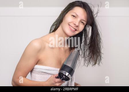 Jolie femme dans une serviette sèche-cheveux bruns avec un sèche-cheveux à main dans sa salle de bain regardant la caméra avec un sourire chaleureux et amical Banque D'Images