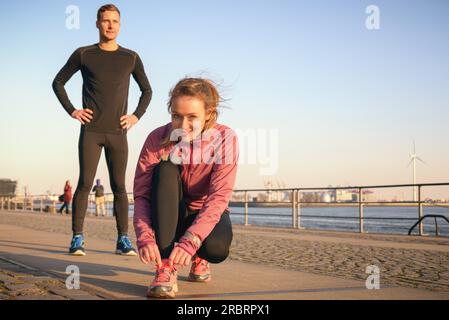 Couple sportif actif sur une promenade en bord de mer se préparant à aller pour un jogging dans leur entraînement quotidien avec la jeune femme attrayante se penchant vers le bas pour attacher Banque D'Images