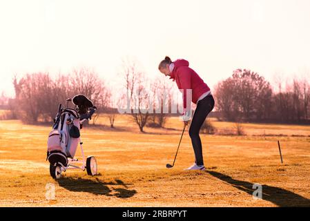 Femme golfeuse jouant une partie de golf dans la soirée de soleil en alignant sur le fairway pour un tir avec sa voiturette de golf au premier plan et long Banque D'Images