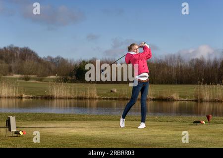 Femme golfeur frappant une balle de golf sur le fairway avec un club debout dans la position de suivi après le coup devant un étang ou de l'eau Banque D'Images