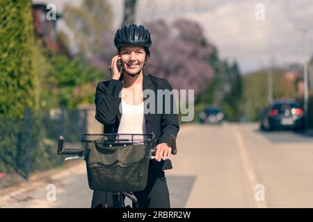 Élégante jeune femme d'affaires amicale se rendant au travail en pause pour répondre à un appel sur son téléphone portable debout soutenant son vélo dans la route comme elle Banque D'Images