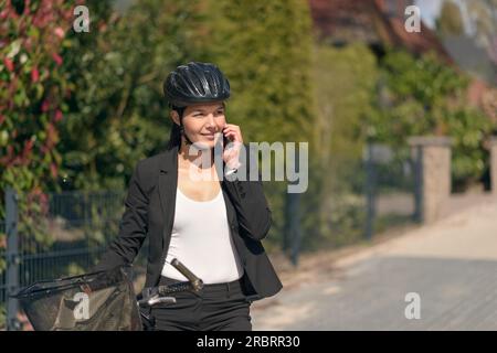 Élégante jeune femme d'affaires amicale se rendant au travail en pause pour répondre à un appel sur son téléphone portable debout soutenant son vélo dans la route comme elle Banque D'Images