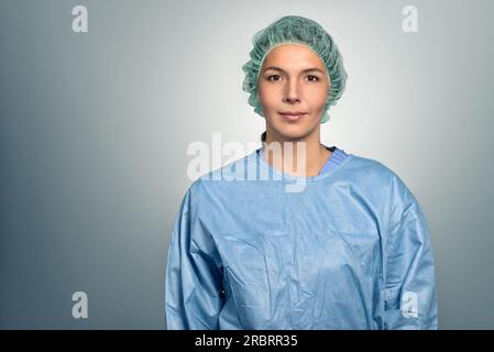 Séduisante femme d'âge moyen médecin ou infirmière en gommage et capuchon de stérilité regardant directement la caméra sur un fond gris, la tête et les épaules Banque D'Images
