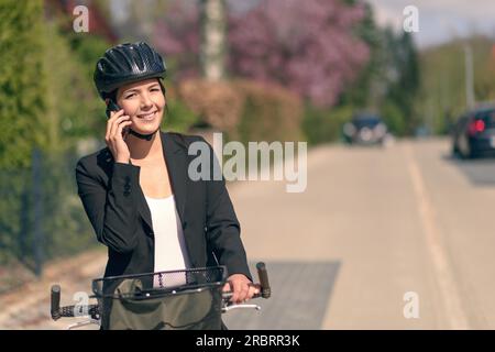 Élégante jeune femme d'affaires amicale se rendant au travail en pause pour répondre à un appel sur son téléphone portable debout soutenant son vélo dans la route comme elle Banque D'Images