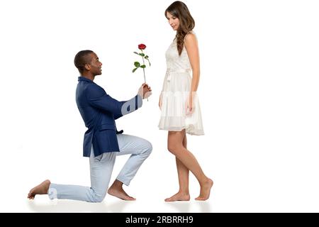 Romantique hipster pieds nus jeune homme agenouillé sur le sol proposant à son amoureux de déclarer son amour éternel dans une scène tendre, couple multiracial Banque D'Images