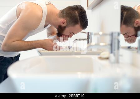Homme rinçant sa brosse à dents sous l'eau courante du robinet dans le lavabo après s'être rasé au-dessus de sa barbe pour neaten son apparence, vue de côté Banque D'Images