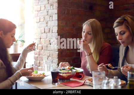 Trois jeunes amies attrayantes se détendant autour du déjeuner ensemble dans un restaurant assis à une table bavardant et appréciant leur nourriture Banque D'Images
