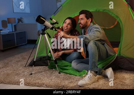 Couple heureux utilisant le télescope pour regarder les étoiles tout en étant assis dans la tente de camping à l'intérieur Banque D'Images