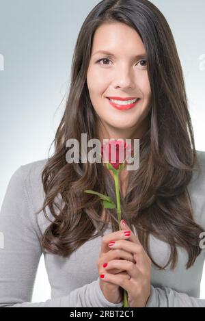 Femme attrayante avec les cheveux longs-bruns tenant une rose rouge à longue tige, un cadeau d'un être cher pour la Saint-Valentin ou l'anniversaire Banque D'Images