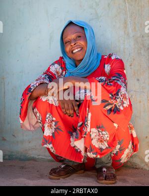 Vêtue de façon vibrante d'une robe écarlate ornée de fleurs, la joyeuse dame tchadienne dans un hijab bleu vif nous honore de son sourire radieux, assise près d'un mur Banque D'Images