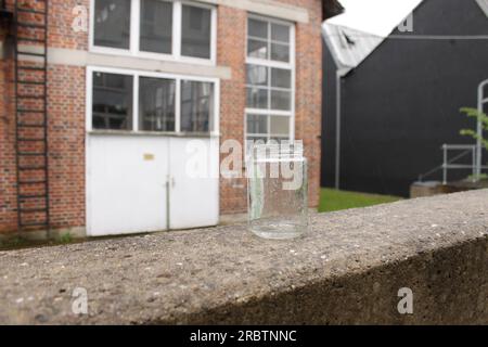 Vieux bocal utilisé comme cendrier couvert de gouttes de pluie en se tenant debout sur un mur de béton. Un bâtiment industriel abandonné en briques en arrière-plan. Banque D'Images