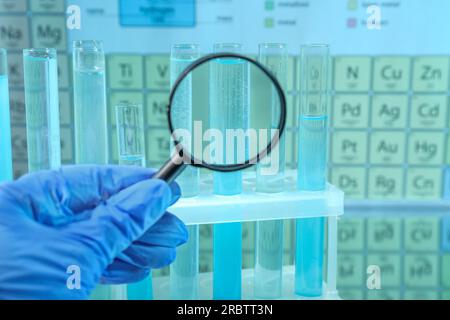 Assistant de laboratoire regardant à travers la loupe sur des tubes à essai contre le tableau périodique des éléments chimiques, gros plan Banque D'Images