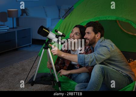 Couple heureux utilisant le télescope pour regarder les étoiles tout en étant assis dans la tente de camping à l'intérieur Banque D'Images