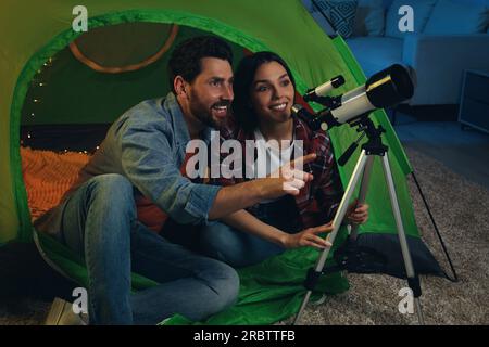 Couple heureux utilisant le télescope pour regarder les étoiles tout en étant assis dans la tente de camping à l'intérieur Banque D'Images