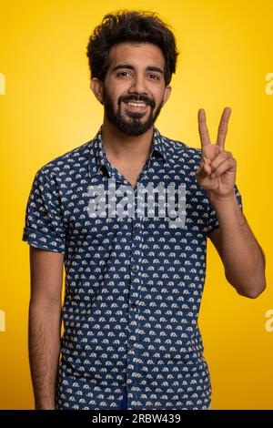 Jeune homme indien montrant le signe de la victoire, espérant le succès et la victoire, faisant geste de paix, souriant avec une expression optimiste gentille. Beau gars hindou barbu isolé seul sur fond de studio jaune Banque D'Images