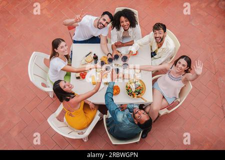 Vue de dessus à angle élevé d'un groupe de meilleurs amis grillant des verres à vin souriant et regardant vers la caméra. Les jeunes adultes qui s'amusent lors d'un dîner sur le toit en prenant des boissons. Réunion de copains. Photo de haute qualité Banque D'Images