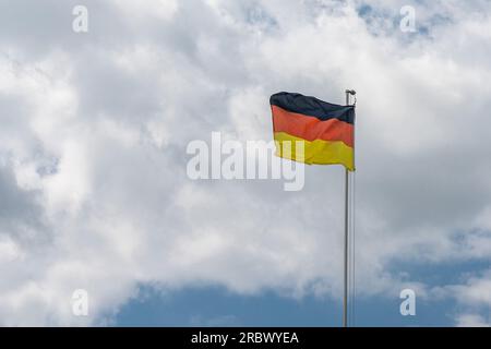 Le drapeau national allemand sur un mât métallique flotte dans le vent contre un ciel nuageux. Banque D'Images