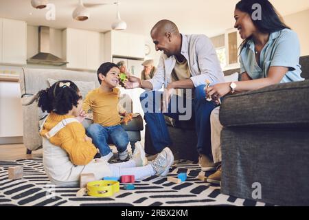 Maison familiale, enfants et parents avec des jouets, salon et ludique sur le sol avec parler, rire et jeux. Hommes, femmes et enfants avec bâtiment Banque D'Images