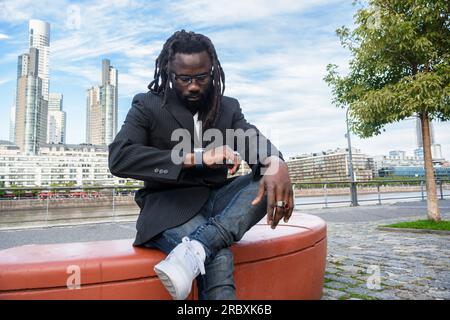 Jeune homme d'affaires africain avec barbe, dreadlocks et lunettes, en vêtements décontractés, porte une veste bleue, attend dehors, assis à regarder l'heure, Banque D'Images