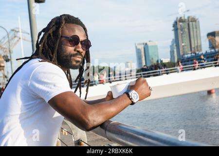 Jeune homme noir avec des dreadlocks, barbe et lunettes de soleil, debout appuyé sur la balustrade appréciant le coucher du soleil dans le quartier de Puerto Madero Banque D'Images