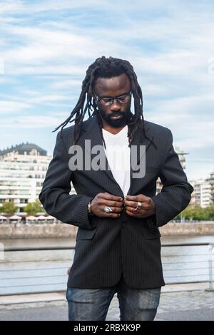 portrait vertical d'un jeune homme d'affaires africain à la mode avec des dreadlocks, barbe et lunettes sombres, debout à l'extérieur boutonnant blazer. Banque D'Images