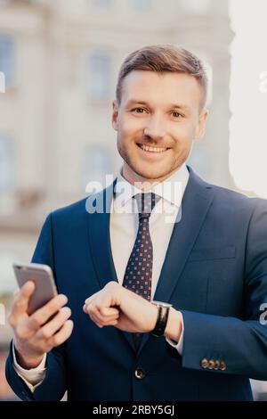 Portrait de beau directeur exécutif masculin bien habillé et joyeux vérifie l'heure sur sa montre-bracelet, utilise un téléphone intelligent, vérifie la boîte e-mail et attend la banque Banque D'Images