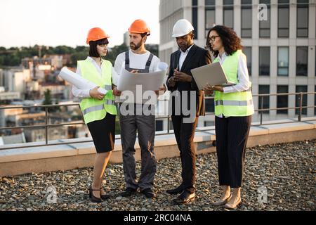 Équipe d'architectes et de designers multiraciaux debout sur la terrasse panoramique et regardant ensemble les schémas du projet. Dame afro-américaine tenant r Banque D'Images