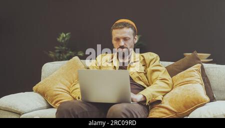 Homme assis sur un canapé avec un ordinateur portable, engagé dans le travail ou d'autres activités numériques. Le réglage détendu et confortable du canapé suggère que l'homme est dans le bureau à domicile ou l'environnement intérieur confortable. . Photo de haute qualité Banque D'Images