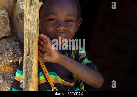 Regardant à travers les yeux rétrécis, un jeune garçon tchadien se tient à l'entrée de sa maison nomade traditionnelle, vêtu d'un T-shirt rayé vibrant Banque D'Images