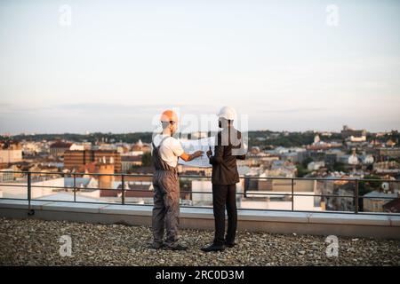 Vue arrière du contremaître en uniforme gris et de l'investisseur en costume noir regardant ensemble les dessins techniques tout en se tenant debout sur la terrasse panoramique. Constructeurs masculins discutant du projet immobilier. Banque D'Images