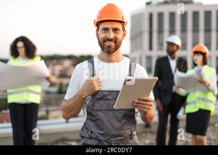 Portrait de contremaître barbu souriant en casque dur et uniforme posant sur la caméra avec tablette numérique dans les mains. Équipe d'investisseurs multiethniques vérifiant le projet de construction tout en se tenant sur la terrasse panoramique. Banque D'Images